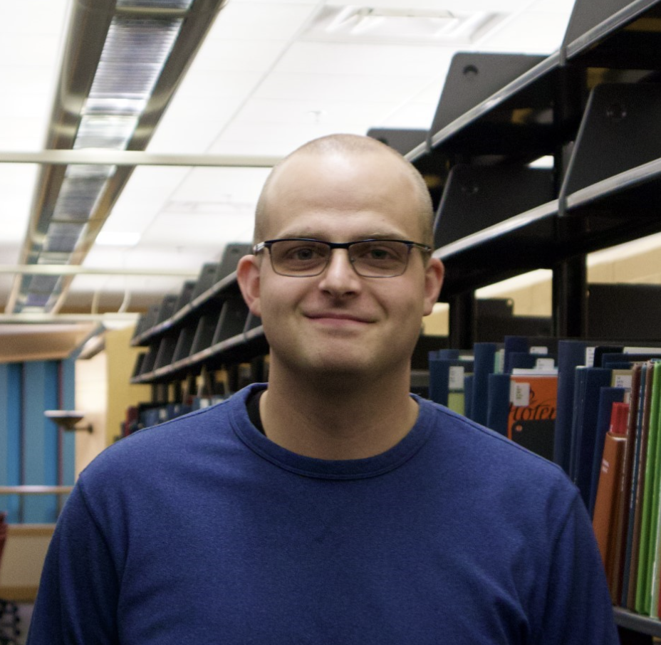 A person with short hair and glasses wearing a blue shirt stands and smiles in a library aisle with bookshelves in the background.
